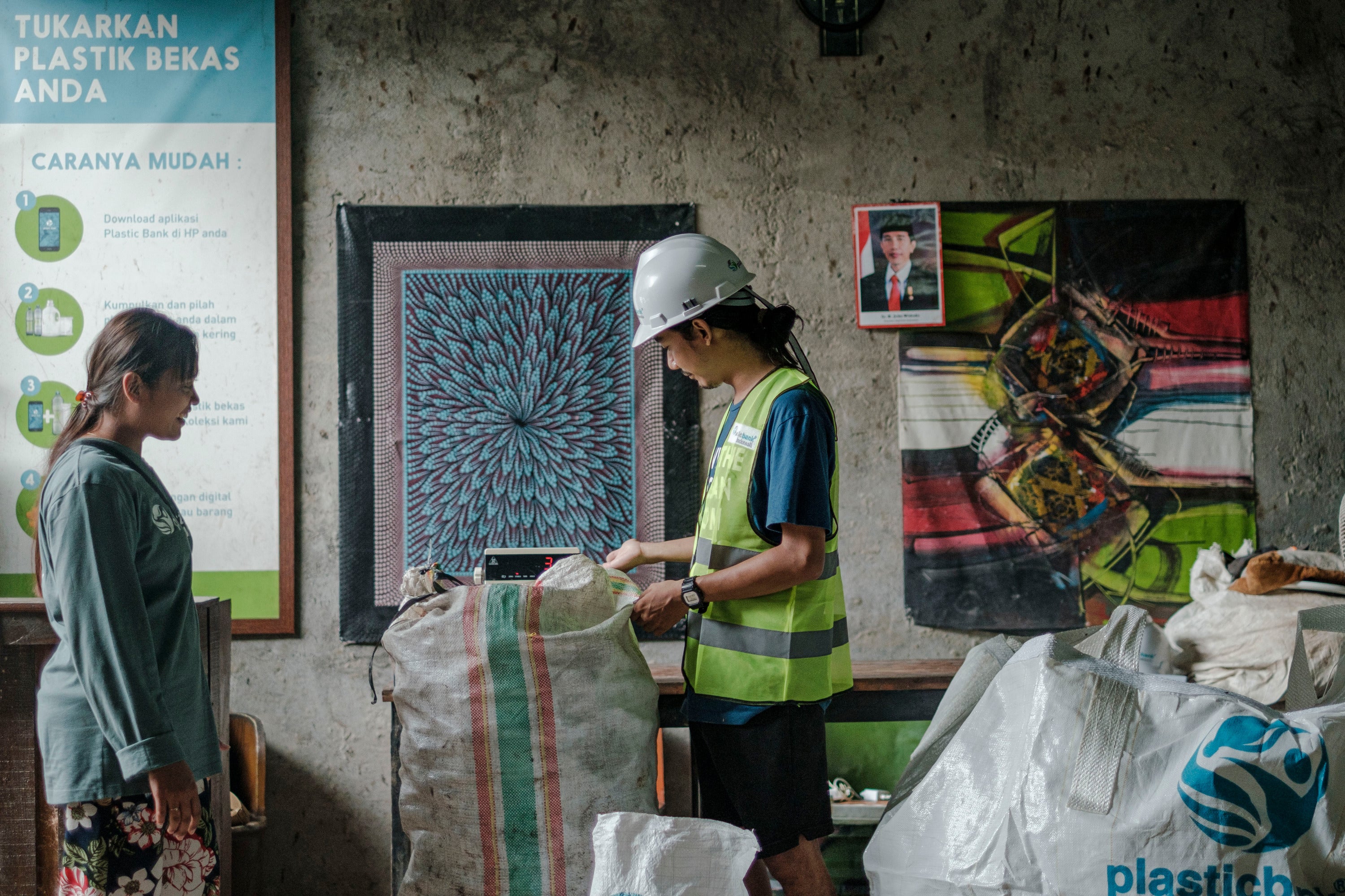 Person in a safety helmet and reflective vest standing next to a woman in a room with artworks on the wall.