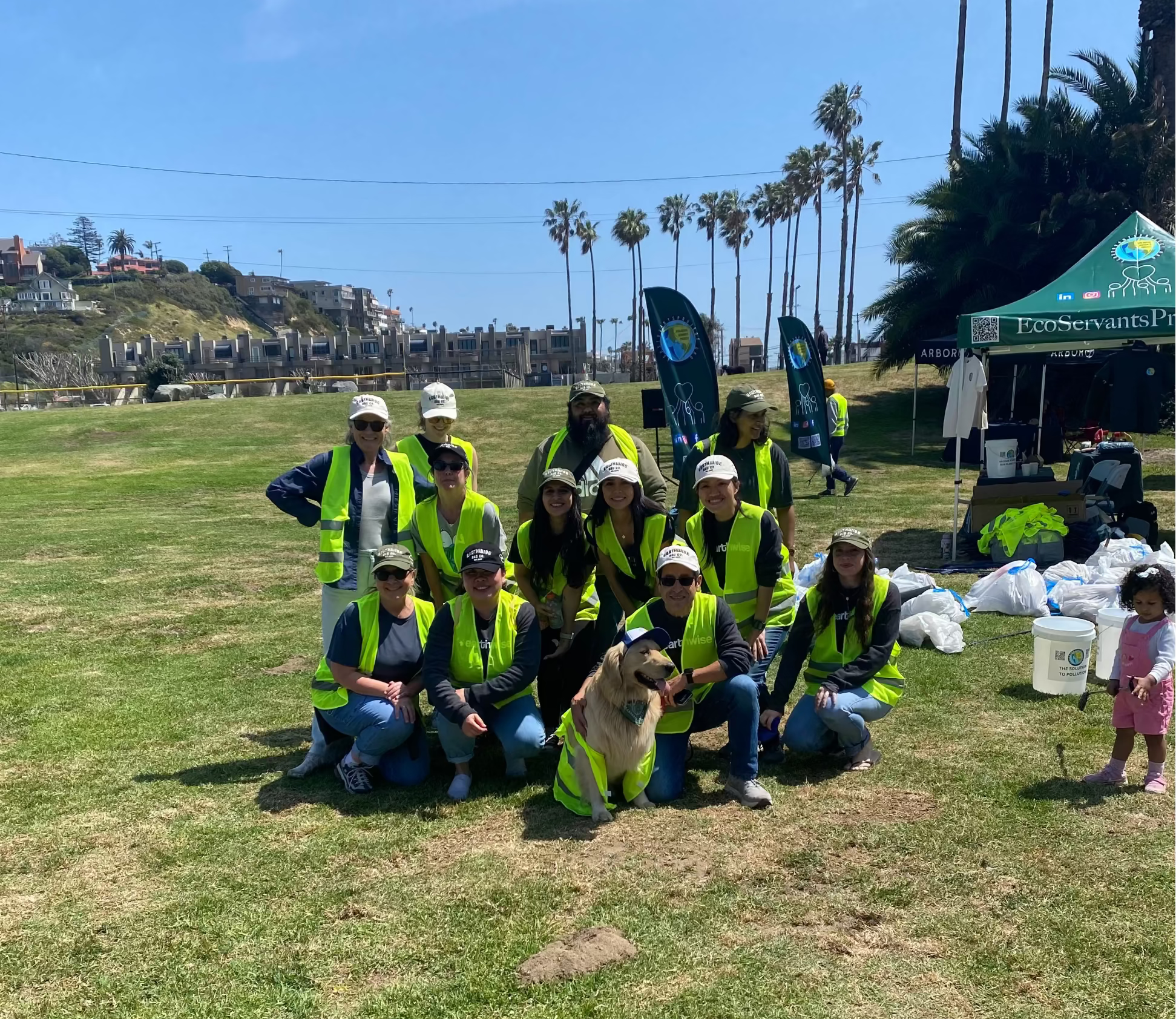Group of people in high-visibility vests posing with a dog on a grassy field, palm trees in the background.